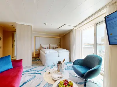 Moderne Suite an Bord der AIDAdiva mit Doppelbett, rotem Sofa und blauem Sessel. Auf dem Tisch sind Champagner und frisches Obst. Große Glastüren bieten Zugang zu einem Balkon.