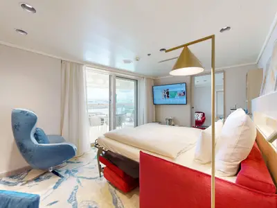 Helle, moderne Suite an Bord der AIDAdiva. Grosses Bett, rotes Sofa, blauer Sessel, Flachbild-TV und Balkon mit Sitzgelegenheiten sowie Hafenblick.