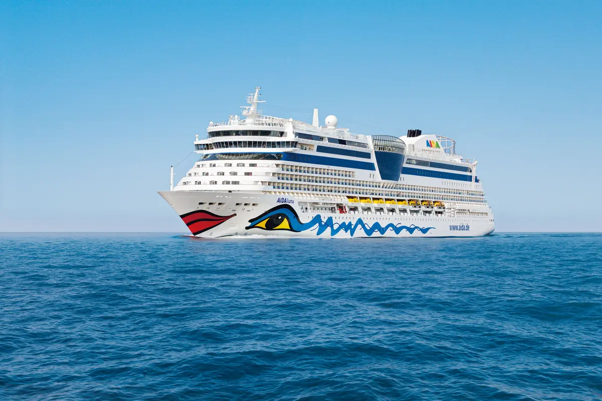 Das Kreuzfahrtschiff AIDAluna ist in einer weiten Einstellung auf dem offenen Meer unter klarem, blauem Himmel zu sehen. Die Front des Schiffes mit dem markanten Kussmund-Design und der Schiffsname AIDAluna sind deutlich erkennbar, während das Schiff nach rechts fährt und sanfte Wellen hinterlässt.