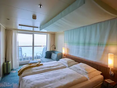 Die geräumige Balkonkabine an Bord der AIDAluna zeigt ein helles Zimmer mit zwei Einzelbetten, einem türkisen Sofa, einem kleinen Beistelltisch und direktem Zugang zum Balkon mit Meerblick. Ein leuchtend grüner Koffer steht neben einem Schrank. Warme Lichter erhellen das Bett.