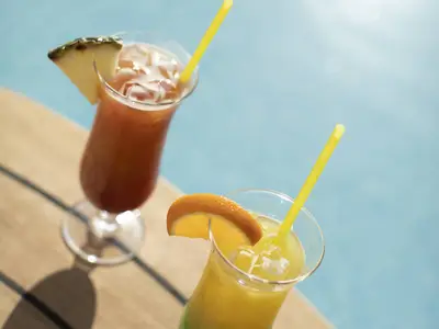Zwei bunte Cocktails, garniert mit Ananas und Orange, stehen auf einem Holztisch neben einem hellblauen Pool an Bord der AIDAluna. Die Getränke enthalten Eiswürfel und gelbe Strohhalme.