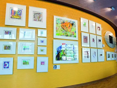 An Bord der AIDAluna: Eine gelbe Wand in der Kunstgalerie, voll mit gerahmten Kunstwerken verschiedener Größen und Stile, darunter bunte Texte und abstrakte Gemälde. Lichtreflexe in den Glasrahmen.