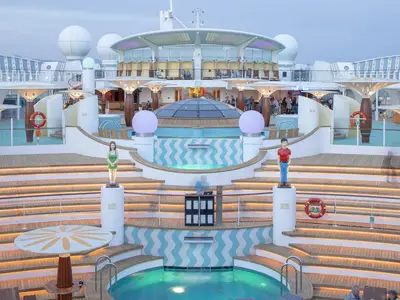 Das abendliche Pooldeck der AIDAluna zeigt eine amphitheaterartige Anordnung mit beleuchteten Stufen, Pools und Bars. Mehrere Figuren stehen auf Podesten, während Gäste im Hintergrund die Atmosphäre genießen.