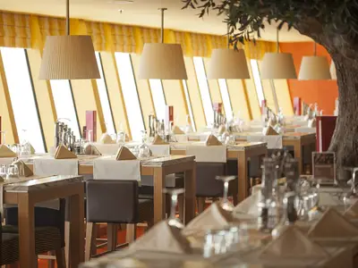 Blick ins Bella Vista Restaurant der AIDAluna. Zahlreiche Tische sind elegant gedeckt mit Gläsern und Servietten. Grosse Fenster und Pendelleuchten erhellen den Raum.