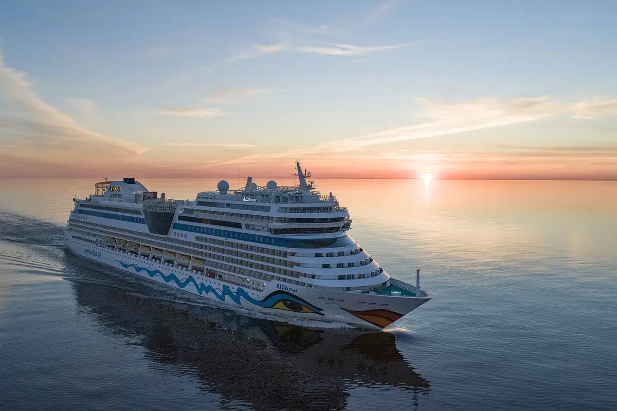Die AIDAmar kreuzt bei Sonnenuntergang auf einem ruhigen Meer. Schiff ist von links nach rechts ausgerichtet, untergehende Sonne spiegelt sich im Wasser.