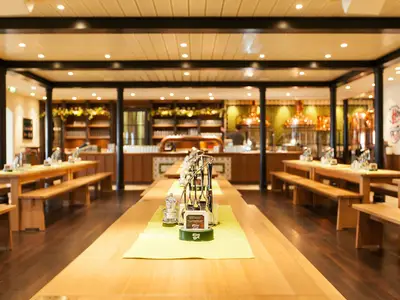 Das helle Brauhaus an Bord der AIDAmar zeigt eine Reihe leerer Holztische mit Bänken und eine Bar im Hintergrund mit Braukesseln. Die Decke ist hell und mit Lichtern versehen.