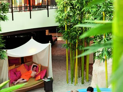 Eine lächelnde Frau liegt entspannt in einem Tagesbett mit bunten Kissen im Spa-Bereich der AIDAmar, umgeben von hohen Bambuspflanzen und einem gefliesten Boden. Sie liest ein Buch.
