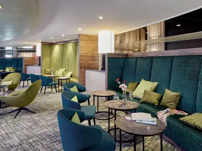 Gemütliche Lounge an Bord der AIDAnova mit grünen Sofabänken, blauen Sesseln und runden Tischen. Ein moderner Teppich und Holzelemente schaffen eine entspannte Atmosphäre.