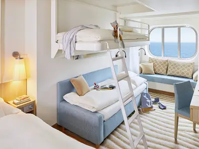 Detailansicht einer modernen Familienkabine mit Meerblick auf der AIDAnova. Man sieht Stockbetten, ein Schlafsofa, einen Schreibtisch und ein ovales Fenster mit Blick auf das Meer.