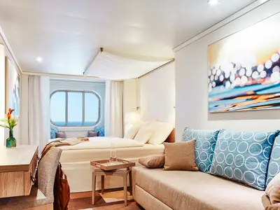 Moderne Meerblickkabine an Bord der AIDAnova. Sie verfuegt ueber ein ovales Fenster mit Meerblick, ein Doppelbett, ein Sofa mit blauen Kissen und einen Schreibtisch.