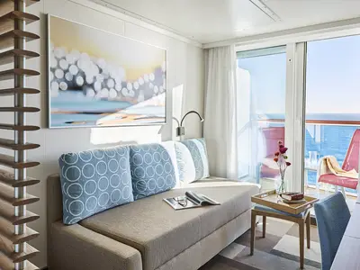 Eine moderne Balkonkabine an Bord der AIDAnova mit einem beigefarbenen Sofa, blauen Kissen und einem privaten Balkon mit Meerblick und rotem Stuhl.
