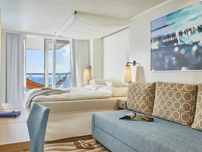 Eine moderne Verandakabine Deluxe an Bord der AIDAnova. Das Schlafzimmer hat ein großes Bett, ein Sofa mit Kissen und einen Balkon mit Hängematte und Meerblick.