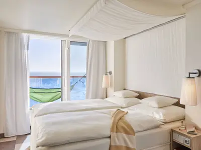 Verandakabine Comfort auf der AIDAnova. Das Schlafzimmer hat zwei Einzelbetten mit weisser Bettwäsche. Der Blick geht auf den Balkon mit Meer und grüner Hängematte.