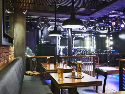 Innenansicht der Rock Box Bar an Bord der AIDAnova. Biere auf Holztischen im Vordergrund. Im Hintergrund eine hell beleuchtete Bühne mit Instrumenten. Links eine Sitzbank mit Ziegelwand.
