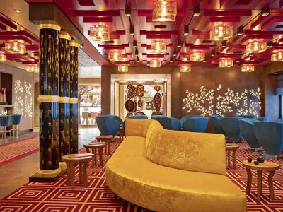 AIDAnova Tokyo Bar: Elegante Lounge mit blauen Sesseln, gelbem Sofa, rotem Teppich. Dunkle Säulen mit Schriftzeichen, goldene Deckenleuchten. Asiatisches Design.