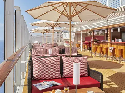 Die einladende Poolbar an Bord der AIDAnova mit bequemen Lounge-Sesseln, Sonnenschirmen und Holztheke mit Barhockern. Im Hintergrund ist das blaue Meer zu sehen.