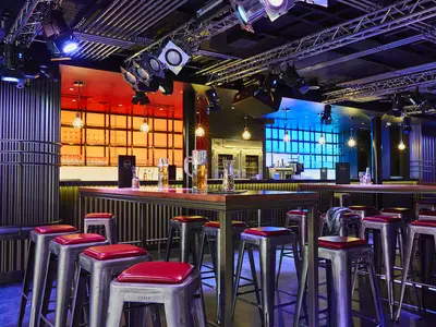 Moderne Bar-Lounge an Bord der AIDAnova. Hohe Tische mit roten Hockern, beleuchtete Bar in Rot und Blau. Deckenkonstruktion mit Bühnenleuchten. Gläser und Getränke sind sichtbar.