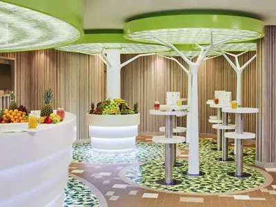 Innenansicht der modernen Juicy's Smoothiebar an Bord der AIDAnova. Man sieht eine Theke und eine Obstinsel mit frischen Früchten, weißen Tischen, grünen Deckenleuchten und Baum-Dekorationen.