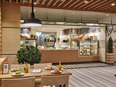 Der moderne Street Food Bereich der AIDAnova zeigt Buffets mit Speisen und Gebäck, Crewmitglieder und Tische mit Gästen. Die Einrichtung ist hell und holzbetont.