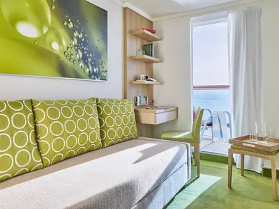 Helle, moderne Balkonkabine an Bord der AIDAnova mit einem Sofa, Schreibtisch und Balkon mit Meerblick. Grüne Kissen und Teppich sorgen für Farbtupfer.