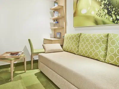 Einzelkabine der AIDAnova. Beiges Sofa mit grünen Kissen, Holztisch, Wandregal mit Schreibtisch. Helle Einrichtung in Grün und Beige.