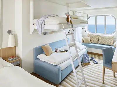 Helle Familienkabine der AIDAnova mit Meerblick. Links ein Einzelbett, mittig ein Schlafsofa mit Hochbett und Leiter. Rechts ein Schreibtisch und eine Fensterbank mit Polstern.