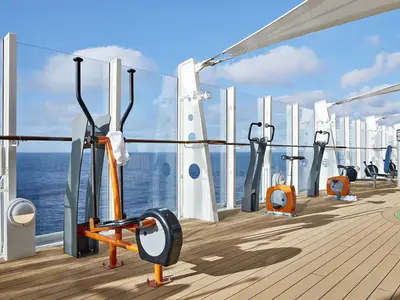 Der Outdoor-Fitnessbereich an Bord der AIDAnova zeigt verschiedene Fitnessgeräte auf einem Holzdeck. Durch eine Glasbrüstung ist das blaue Meer und der bewölkte Himmel zu sehen.