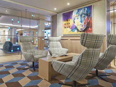 Moderne Lounge an Bord der AIDAnova. Ein farbenfrohes Paul McCartney-Gemälde, blaue Lichtkugeln und karierte Sessel prägen den Raum. Ein geometrischer Teppich in Blau und Braun bedeckt den Boden.