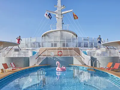 Sonniges Pooldeck der AIDAnova mit grossem Pool und rosa Flamingo. Holzdecks, Liegestühle, Treppen, Skulpturen und Schiffsmast sind sichtbar.