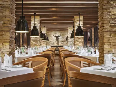 Blick in das Churrascaria Steakhouse an Bord der AIDAnova. Zwei Reihen elegant gedeckter Tische mit Holzsesseln fuehren zu einem Kamin mit Stierschaedel an der Steinwand.