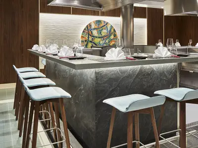 Einrichtung des Teppanyaki Asia Grill Restaurants an Bord der AIDAnova. Ein langer Tresen mit gedeckten Plaetzen und eleganten Hockern in hellem Blau sind zu sehen. Die Theke ist mit einer Unterbeleuchtung versehen, im Hintergrund ist eine farbenfrohe Kunstinstallation. Keine Personen sichtbar.