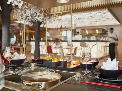 Ein modernes japanisches Restaurant an Bord der AIDAnova. Ein gedeckter Tisch mit Sushi, Schalen und Kochplatte. Im Hintergrund weitere Tische, eine Bar und Personal, umgeben von Kirschbäumen.