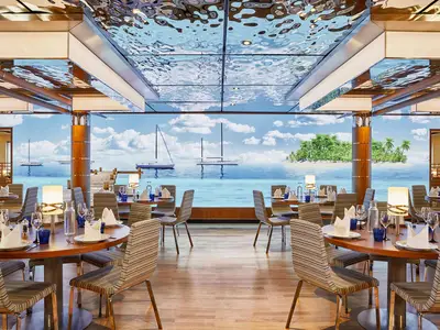 Yachtclub Restaurant an Bord der AIDAnova. Gedeckte Tische und Stühle. Eine Wandprojektion zeigt tropisches Meer mit Yachten und Palmeninsel. Wellenförmige Decke.