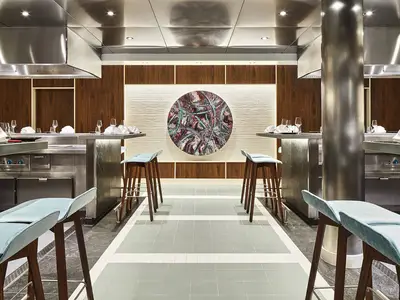 Ansicht des Teppanyaki Asia Grill Restaurants an Bord der AIDAnova mit Kochstationen und Barhockern. Ein abstraktes, rundes Kunstwerk ziert die Wand.