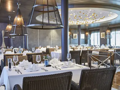 Das elegante Fischrestaurant Oceans an Bord der AIDAnova zeigt stilvoll gedeckte Tische. Moderne Leuchten und eine kunstvolle Deckeninstallation prägen das Ambiente. Im Hintergrund sind Fenster sichtbar.
