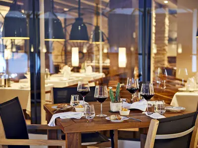 Ein stilvoller gedeckter Holztisch im Restaurant Churrascaria an Bord der AIDAnova. Auf dem Tisch stehen Weingläser, Wassergläser, Geschirr mit Brot und Dips sowie ein kleiner Kaktus. Im Hintergrund sind durch eine Glasfassade weitere Tische und die warme Beleuchtung des Restaurants zu sehen.