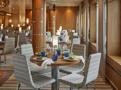 Elegantes Restaurant der AIDAnova. Holzelemente, gestreifte Stühle. Tische mit Geschirr, Gläsern, Salaten gedeckt. Tischlampen schaffen gemütliche Atmosphäre. Ohne Personen.