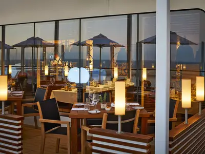 Restaurant an Bord der AIDAnova. Gedeckte Holztische, warmes Licht von Lampen. Große Fenster zeigen Meerblick, Hafen und Schirme im Sonnenuntergang. Ohne Personen.