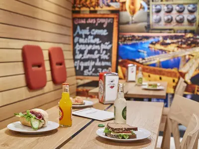 Gemütliches Streetfood-Restaurant auf AIDAnova. Holztische mit Sandwiches (Salat, Gurke, Wurst) und Limonadenflaschen. Im Hintergrund eine Kreidetafel und eine Wand mit Stadtansicht.