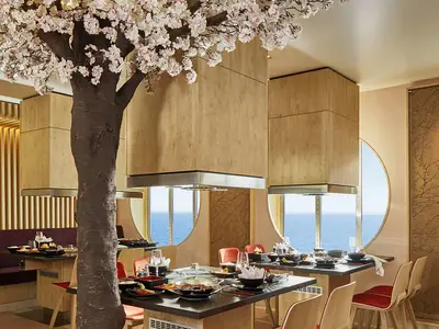 Elegantes japanisches Restaurant an Bord der AIDAnova. Im Zentrum steht ein großer Kirschblütenbaum. Gedeckte Tische und runde Fenster mit Meerblick schaffen eine stilvolle Atmosphäre.
