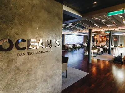 Das 'Ocean's - Das Fischrestaurant' an Bord der AIDAnova. Links das spiegelnde Logo. Rechts der moderne Essbereich mit Holzboden und maritimer Deckeninstallation.