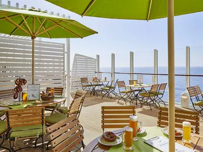Outdoor-Restaurant Brauhaus der AIDAnova. Holztische, Stühle mit grünen Polstern, Sonnenschirme. Bierkrüge und Brezeln auf den Tischen. Blick auf das weite, blaue Meer unter klarem Himmel.