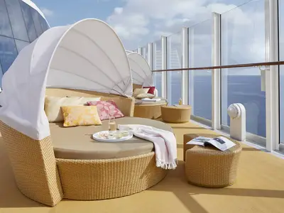 Blick auf das Sonnendeck der AIDAnova. Ein Rattan-Tagesbett mit Kissen und Sonnenschutz, dazu Getränke und ein Buch. Im Hintergrund weitere Betten und das weite, blaue Meer.