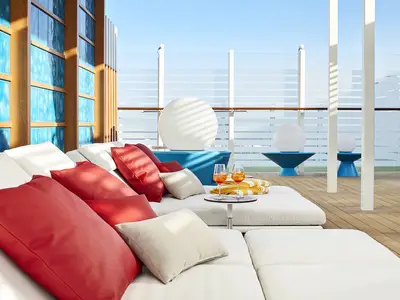 Entspannter Loungebereich an Deck der AIDAnova. Weisse Liegen mit roten Kissen und zwei Cocktails. Blaue Designelemente und gläserne Reling im Hintergrund unter blauem Himmel.