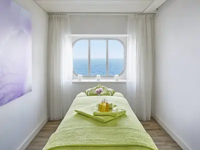 Heller Spa-Behandlungsraum an Bord der AIDAnova. Eine Massageliege mit gruenen Handtuechern, Massageoele und Kerzen. Durch ein ovales Fenster blickt man auf das blaue Meer.