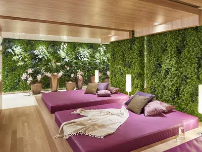 Eine entspannte Wellnessoase an Bord der AIDAnova. Zwei magenta-farbene Lounge-Liegen mit Kissen stehen vor einer grünen Wand aus Pflanzen. Mehrere Stehlampen erhellen den Raum.