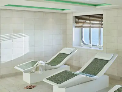 Zwei moderne Heizliegen mit grünen Mosaikauflagen im Spa-Bereich der AIDAnova. Ein Bullauge bietet Meerblick, links die Wand mit Blinds-Schatten.