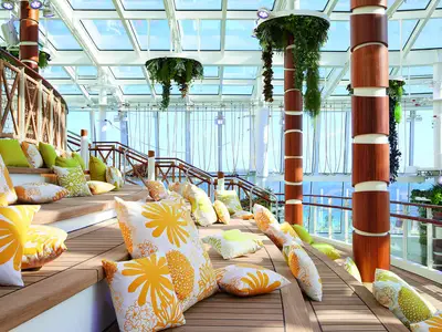 Ein heller, mehrstufiger Lounge-Bereich an Bord der AIDAnova. Bunte Kissen liegen auf Holzstufen unter einem Glasdach. Pflanzen schmuecken Saeulen und Decke. Blick auf das Meer im Hintergrund.