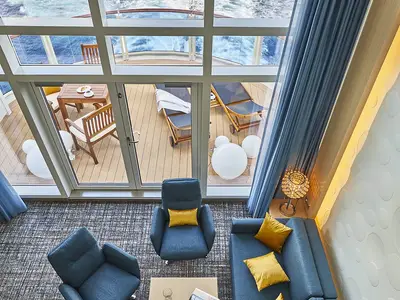 Vogelperspektive in eine moderne Penthouse Suite der AIDAnova. Wohnbereich mit blauen Sesseln und Sofa. Balkon mit Holzmoebeln, Liegestuehlen und weitem Meerblick.