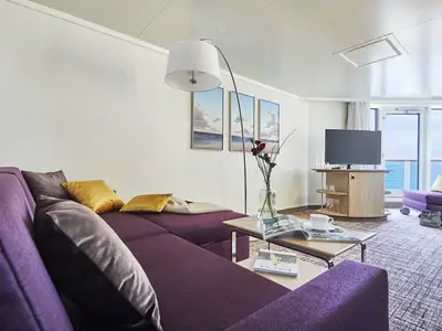 Blick in eine helle, modern gestaltete Suite an Bord der AIDAnova. Ein großes lila Ecksofa mit goldenen Kissen dominiert den Raum, daneben ein Fernseher und ein Balkon mit Meerblick.